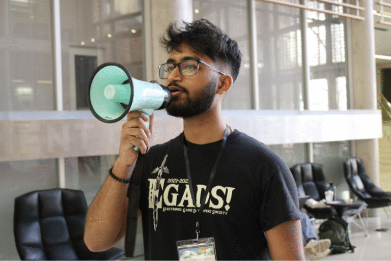 A person wearing glasses and a black “EGADS! Electronic Game Developers Society” T-shirt speaks into a light blue megaphone inside a modern building with tall windows and black lounge chairs in the background.
